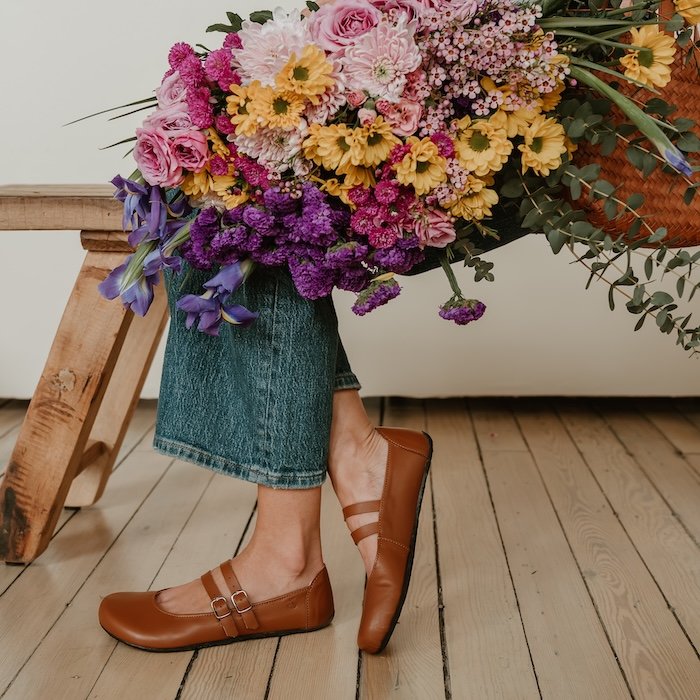 A side view of a woman sitting on a wooden bench. She is visible from the waist down, with a large floral bouquet on her lap, and a pair of Jenon Leather Villet two strap mary janes in a medium brown leather. The right foot is kicked back and resting on the toe behind the left foot.