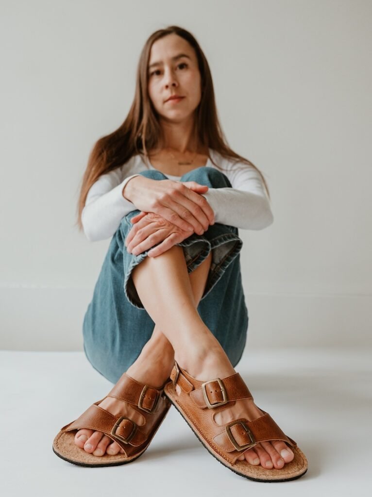 A woman sitting on the ground with her feet crossed in front of her and her hands resting on her knees, she is not smiling, and on her feet are a pair of double buckle Koel Ada leather barefoot sandals.