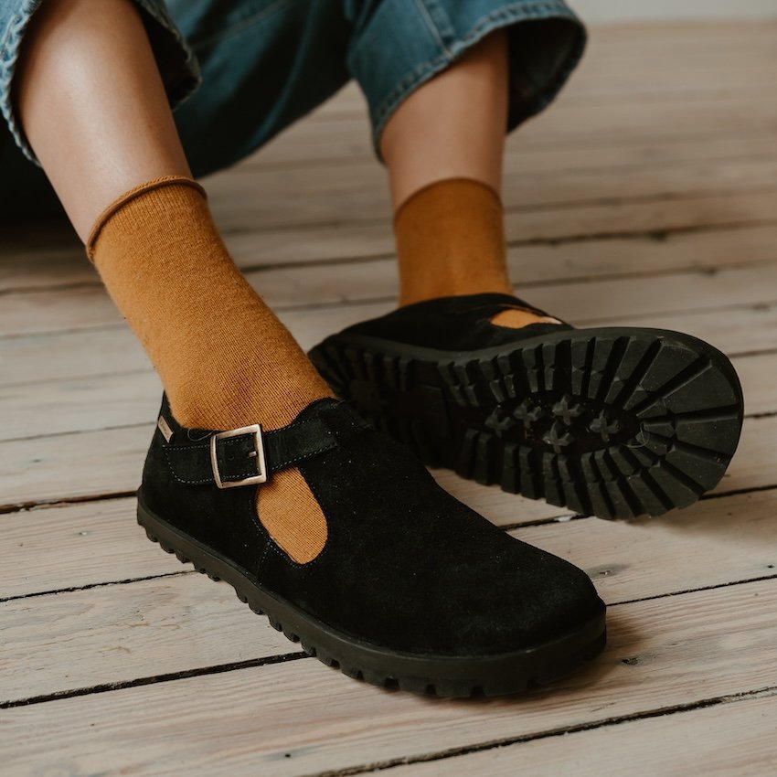 A person's feet close up in a pair of Lang S Mira black suede T strap mary janes, with dark golden socks. The chunky outsole of the left foot is visible and highlighting the wide toe box
