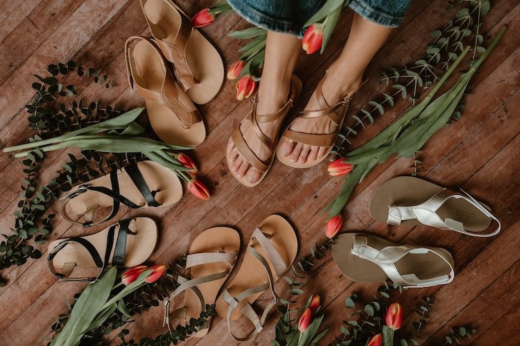 A top down look at a whole collection of sandals from Lang. S by Tikki Shoes showing multiple wide toe box feminine sandal options in a variety of neutrals. They are scattered with flowers.