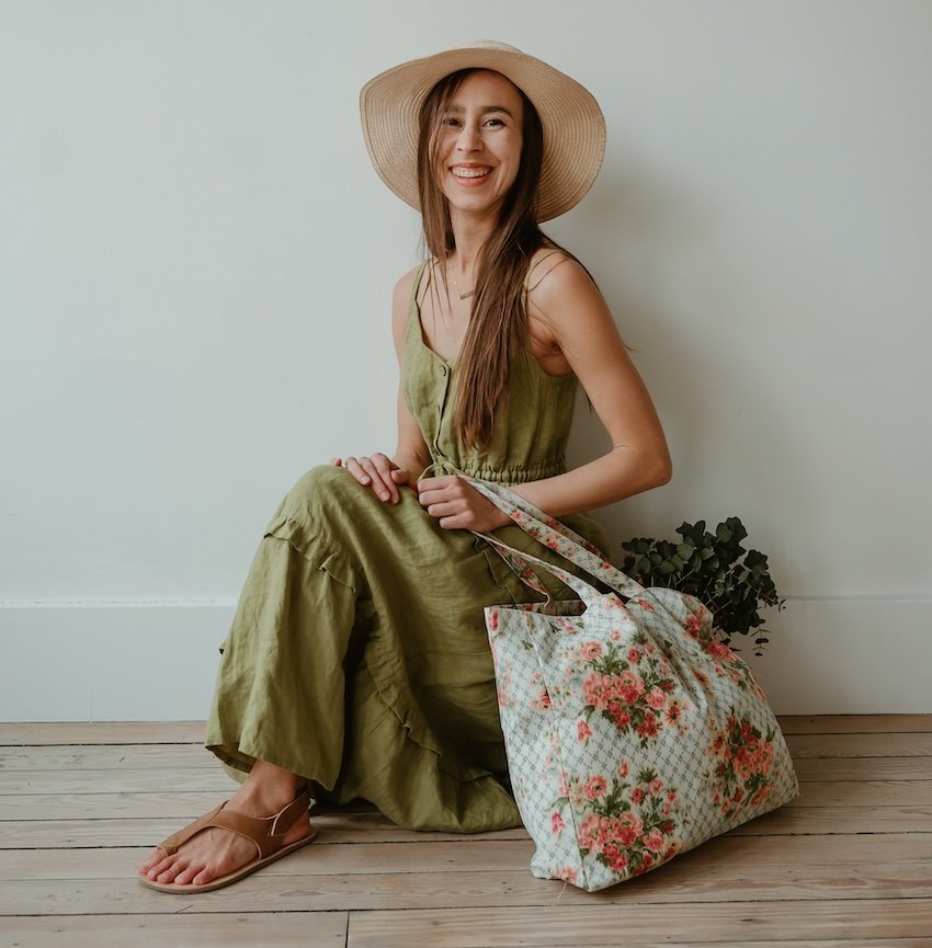 A woman in a light green linen dress and straw hat sitting with a floral bag next to her. She is laughing at the camera, and one foot is showing below her dress, wearing a pair of Lang. S barefoot Sandals in brown leather.