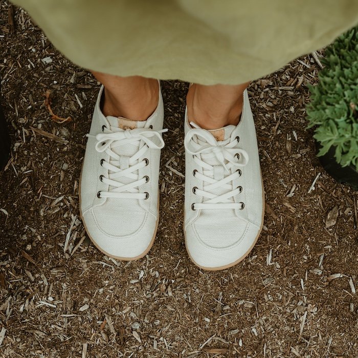 Top down view of a pair of Mukishoes Sand lace up low top barefoot sneakers made with natural sustainable materials. They light colored wide toe box sneakers are being worn on a dirt and wood chip path.