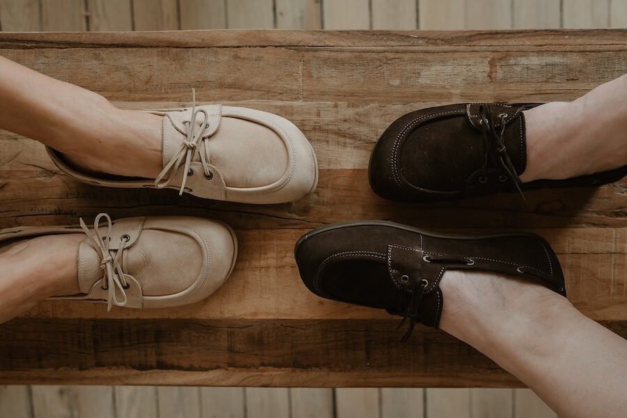 Two people's feet in Peerko Tala resting on a bench. The boat style barefoot shoes are shown in a tan and dark brown with details like leather laces and metal eyelets.