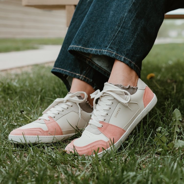 A close up of a person's feet in Shapen ReWind soft peach barefoot sneakers being worn in the grass, with the legs of a pair of dark jeans hanging near the ankles. There are taupe ruffled socks visible just above the sneaker and the flat outsole is shown by the foot being lifted.