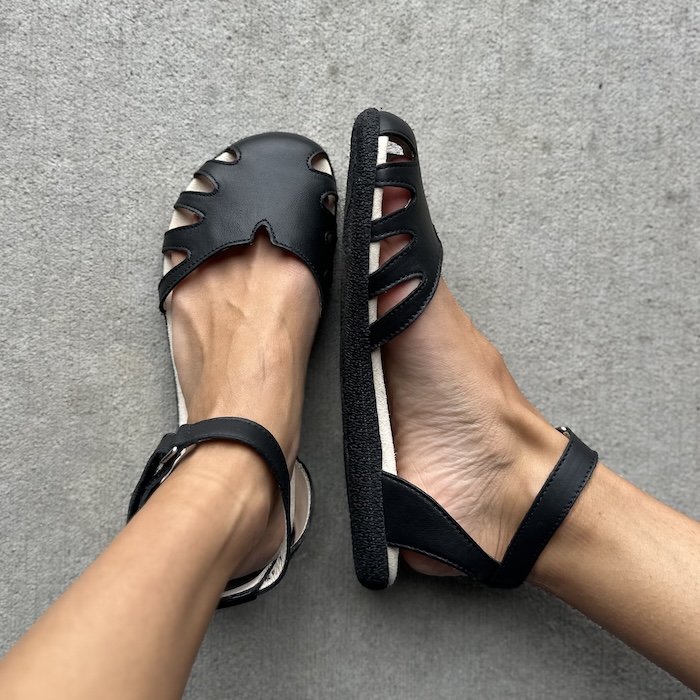 A top down view of a pair of feet resting on concrete wearing black leather Baer Shoes women's dress sandals with a wide toe box