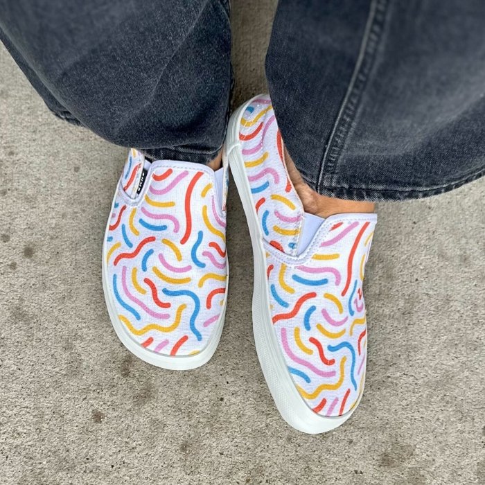 Top down view of a pair of feet in Bohempia Velik Candy barefoot sneakers with brightly colored squiggles on the hemp canvas. The person is wearing a pair of black wide leg jeans and has their left tilted slightly to the side