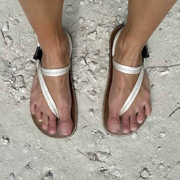 Top down view of a person's feet in Earth Runner's wide toe box sandals in cloud white laces standing on a sandy rocky ground.