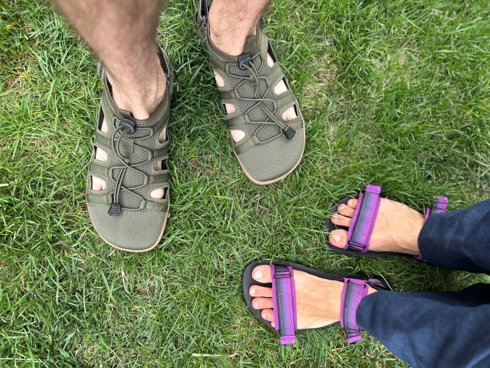 A birds eye view photo of 2 people standing on grass wearing Freet Barefoot sandals. The woman on the right is wearing Pink and Gray Freet Lundy sandals, and the man on the left is wearing Freet Zennor closed toe barefoot sandals in olive green.