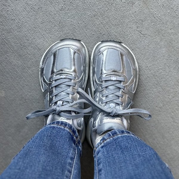 A top down view of a pair of feet standing on concrete visible from the calves down wearing jeans and silver wide toe box Retro Run Ohne Project sneakers