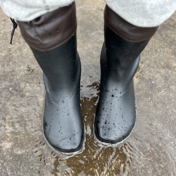 A top down view of a pair of black rubber Saguaro Brisk wide toe box barefoot rain boots, on a person's feet standing in a puddle of water