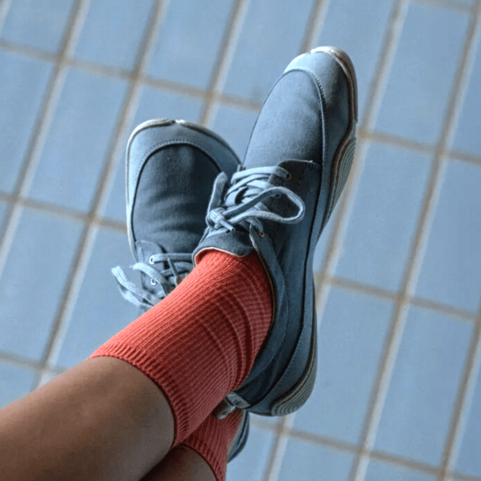 A close up of a pair of crossed feet wearing ribbed orange socks and blue denim-look Wildling Cotinga Petrol barefoot sneakers with a super minimal outsole.