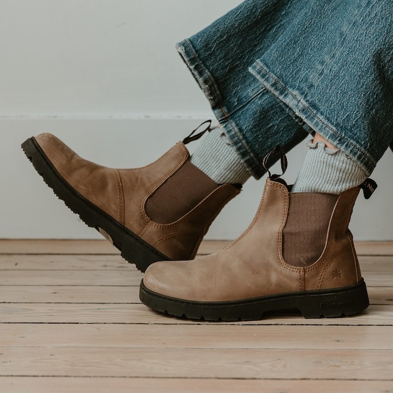 A close up side view of a pair of feet visible from the ankles down wearing jeans, socks, and taupe colored leather El Naturalista Nomada wide toe box chelsea boots with a cushioned sole, resting on a wood floor.