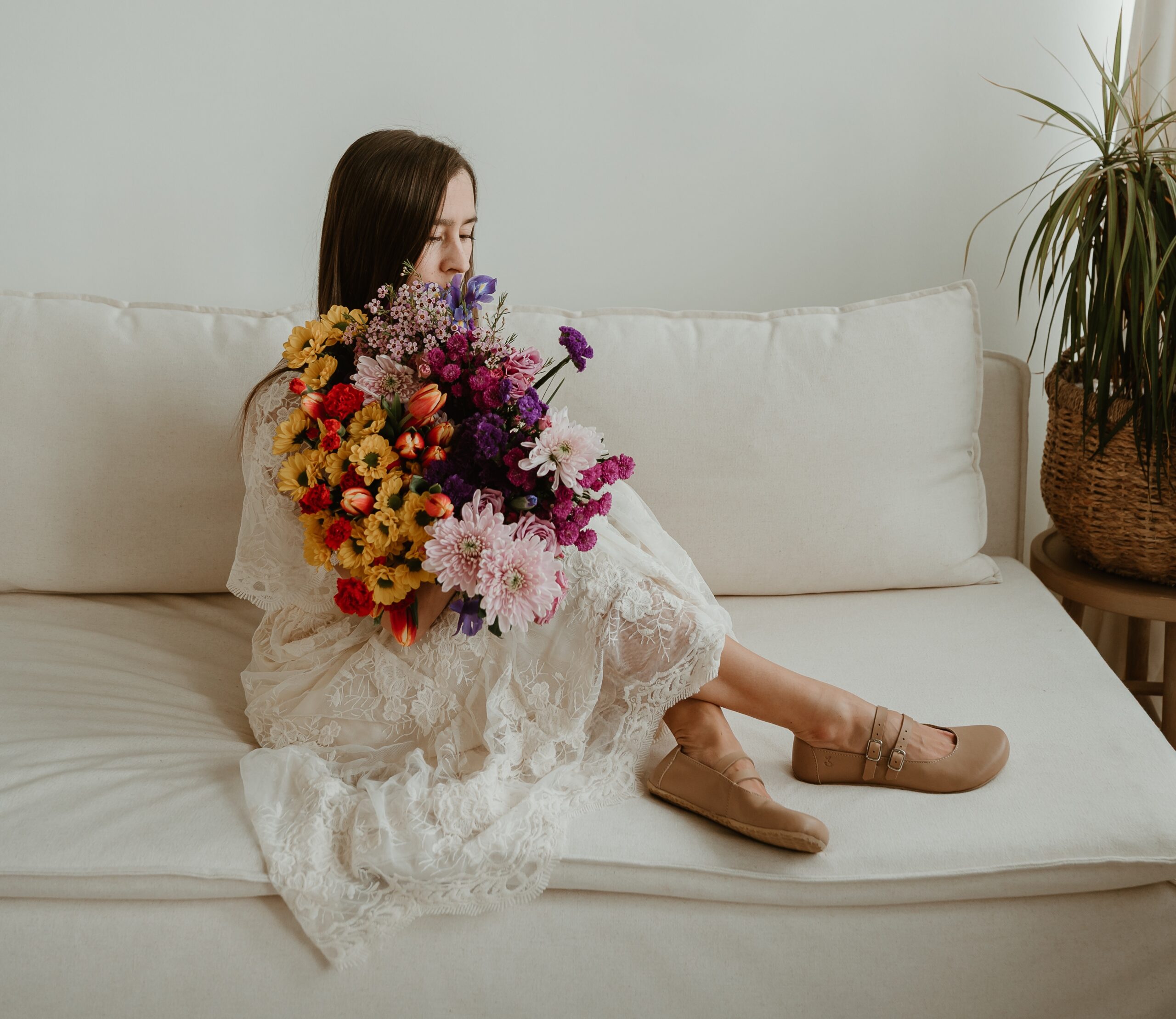 A woman sitting on a white couch in a white lace dress. A large color bouquet of flower is partially covering her face. her feet are crossed in front of her, and she has on a pair of Jenon Leather Villet double strap mary janes in a neutral sand color.