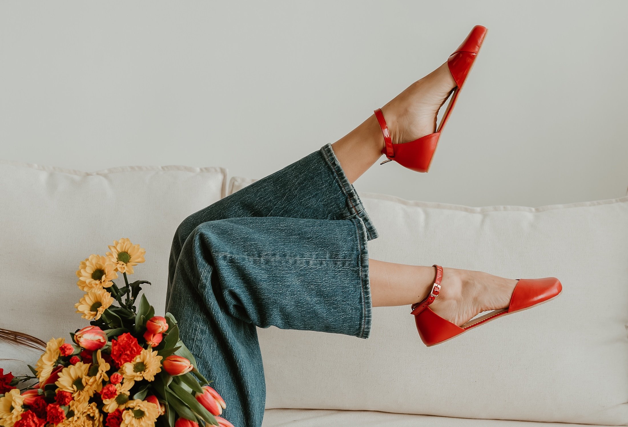 A person's legs in the air in a scissor kick wearing red Shapen Poppy, a barefoot replacement for high heels with a wide anatomical toe box. The person is wearing jeans and holding a colorful bouquet of flower at her waist.
