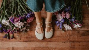 A top down view of a pair of Shapen Orchid in beige suede surrounded by flowers in purple, pink, and raspberry tones. The person is visible from the knees down and the shoes feature a delicate diagonal strap across the foot and a wide toe box.