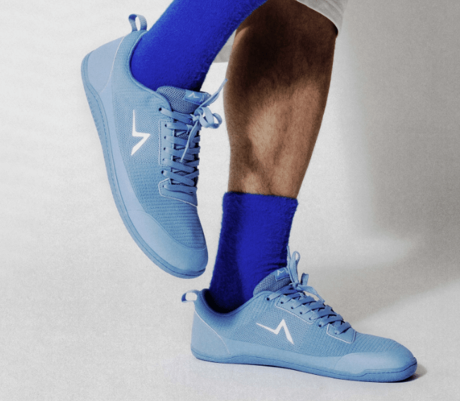 A pair of blue Aion Barefoot training sneakers paired with blue socks being worn by a man. The right foot is lifted and the left foot is flat on the ground.