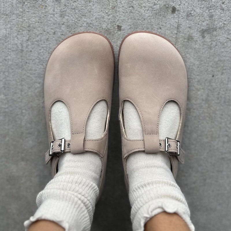 A person's feet in Baer wide toe box t-bar flats with a silver buckle, being worn with Tabio ruffle socks from Anya's Shop. The lighter beige shoes feature a wide toe box and supportive outsole.