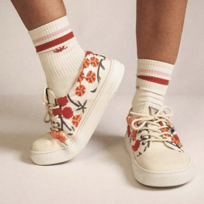 Front view of a person standing in Dolfie Paradise Dylan Embroidered barefoot sneakers with one foot pointing to the side flexed up on the toes. The cream sneakers are embroidered with red and yellow flowers