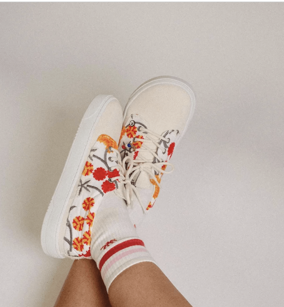 A person's crossed feet in Dolfie Paradise Embroidered barefoot sneakers with pretty red and orange flowers on a cream background
