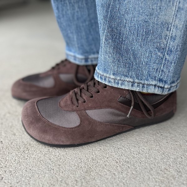 Side view of a pair of Libertas Barellina brown suede and mesh ballet inspired sneaker with a wide toe box and flat flexible outsole. A pair of lighter toned jeans are hanging at the top of the shoes.