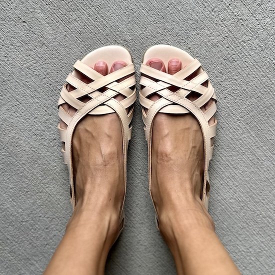 A top down view of a pair of feet resting on concrete wearing Shapen Barefoot Camille natural leather barefoot ballet flats that have an open weave design with delicate straps and a peep toe. They are foot-shaped feminine dress flats.