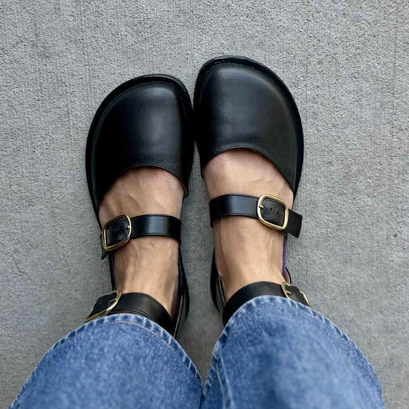 Top close up view of a woman's feet in Shapen Barefoot Neeja black leather flats with buckle details over the foot and around the ankle. Mid tone denim hangs just above the ankle strap.