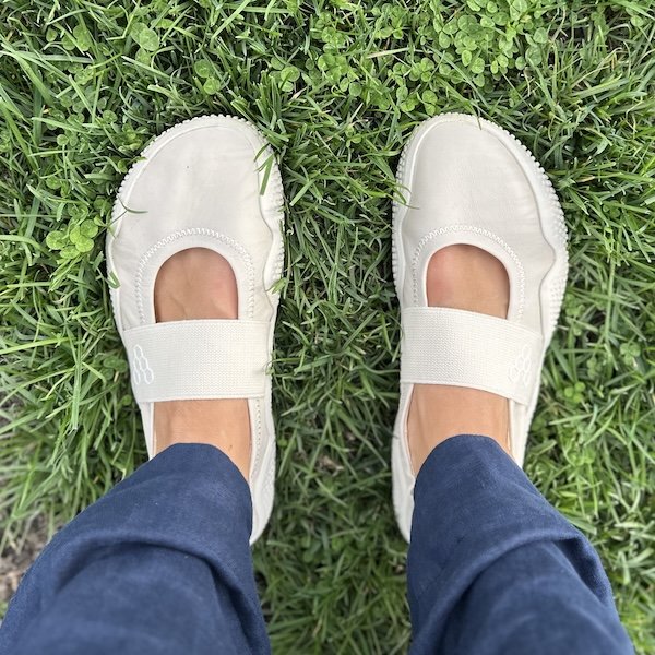 A top down view of a pair of feet standing on grass wearing white leather Vivobarefoot Motus Studio Ballet barefoot flats with a wide anatomical shape to them and and elastic strap