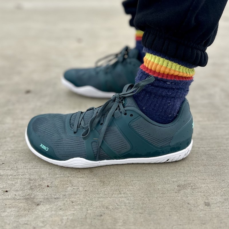 A close up side view of a pair of feet standing on pavement wearing Xero Shoes 360 Rally barefoot court shoes in hunter green