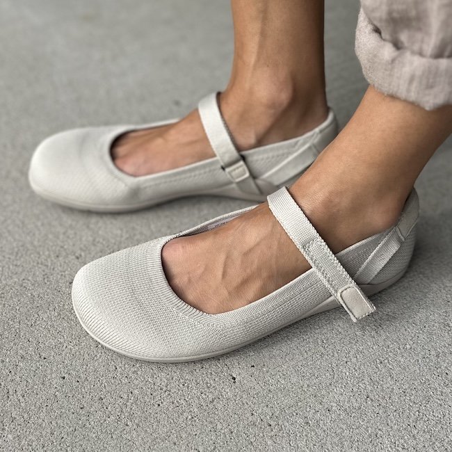 A close up side view of a pair of feet standing on pavement wearing Xero Shoes Knit Cassie barefoot mary jane in beige