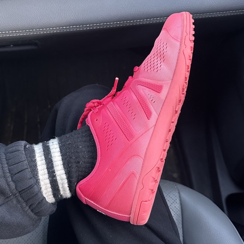A close up of a person with their foot crossed over their leg and the shoe visible. The shoe is a Xero Shoes HFS II women's barefoot athletic sneaker in Raspberry pink