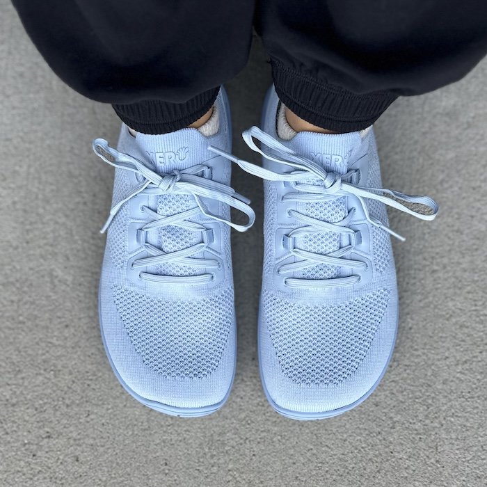 A close up top down view of a pair of feet standing on pavement wearing Xero Shoes Nexus Knit athletic barefoot sneakers in sky blue