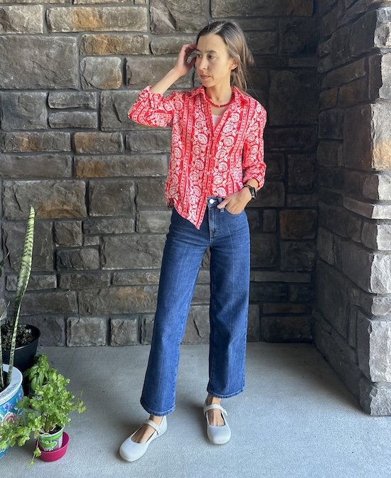A woman standing in a stone entryway wearing a bright floral button down shirt, jeans, and Xero Shoes Cassie Knit barefoot mary jane flats in beige color.
