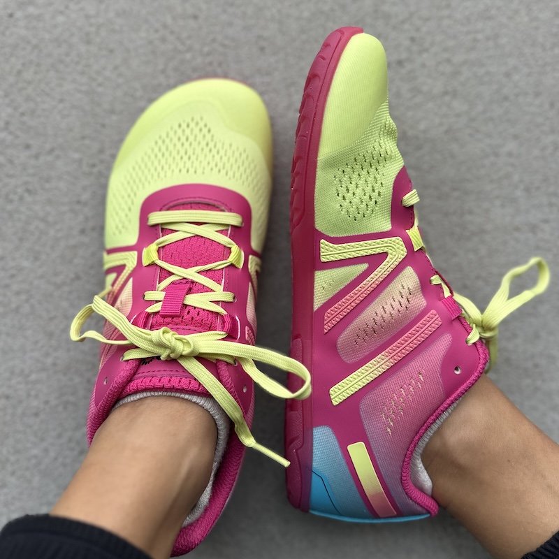 A close up view of a pair of feet resting on pavement wearing Xero Shoes HFS I athletic barefoot sneakers in multicolored mesh