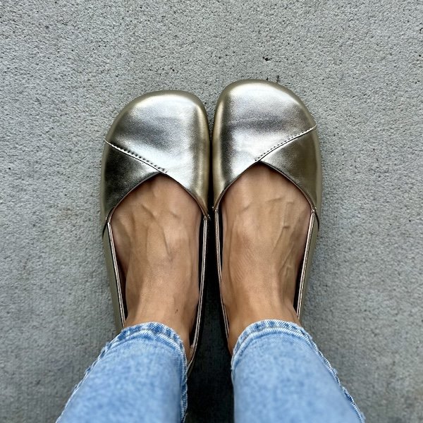 A close up top down view of a pair of feet standing on pavement wearing Xero Shoes Phoenix Leather barefoot ballet flats in gold