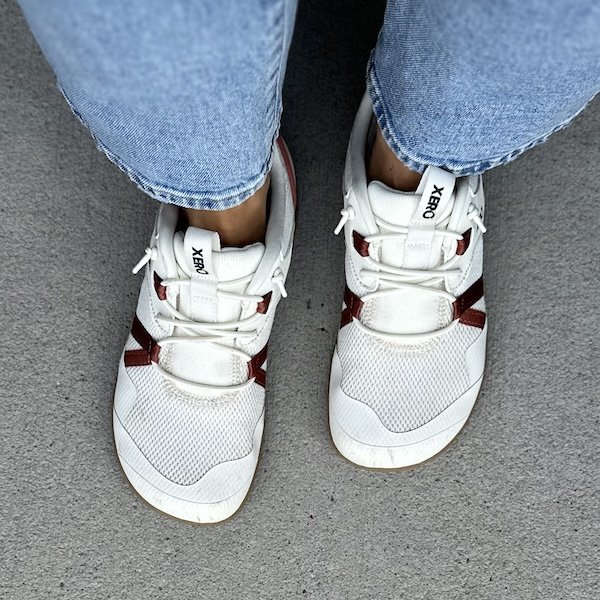 A close up top down view of a pair of feet standing on pavement wearing white Xero Shoes Prio Coast hands free step in shoes