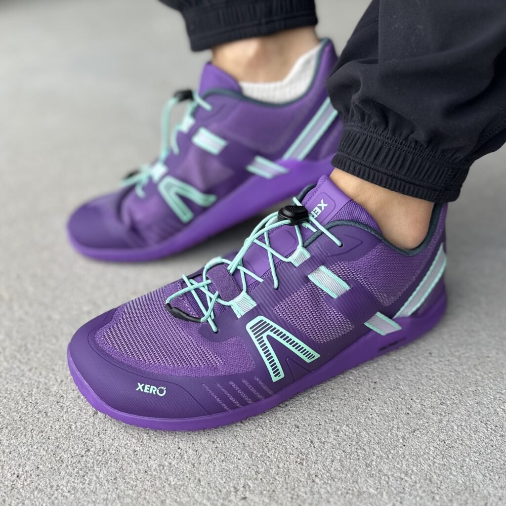 A close up side view of a pair of feet standing on pavement wearing Xero Shoes XT Max barefoot training shoes in vibrant purple