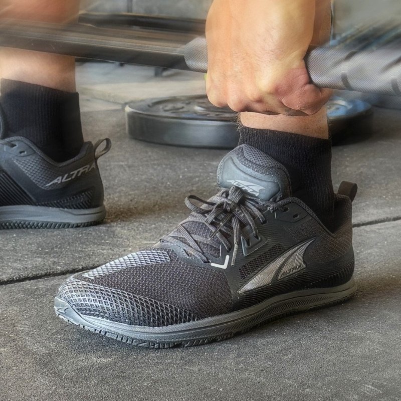 Close up front side look at a pair of Altra Solstice XT 3 wide toe box gym shoe being worn by a person lifting weight in a gym. The all black athletic sneaker is zero drop with a wide toe box