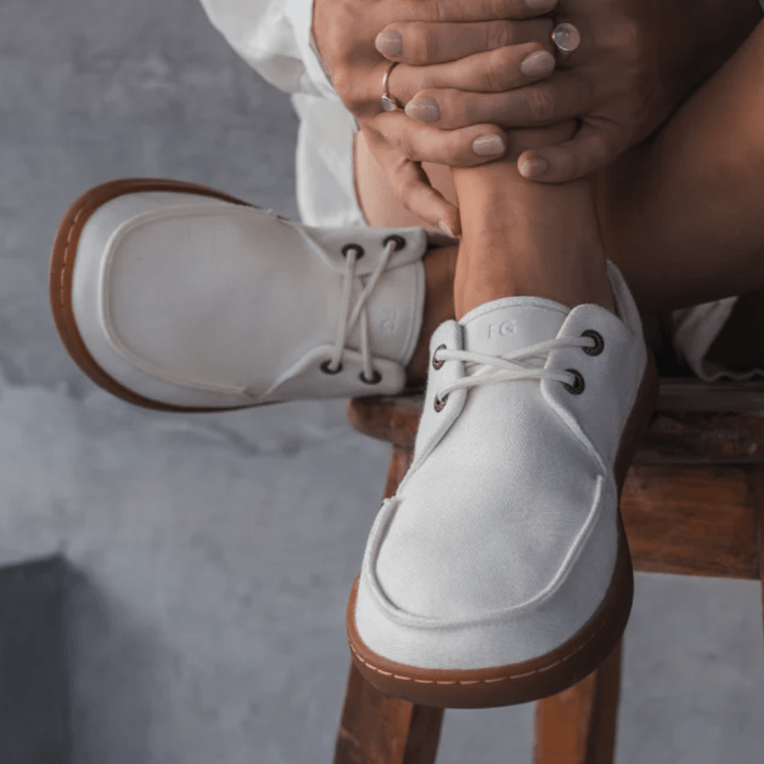 A person sitting on a wooden stool with their legs and feet resting on the seat. hands are held around one leg and on their feet are a pair of Feelgrounds Docks, a canvas barefoot boat shoe with a wide toe box.