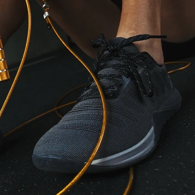 A close up of a person's foot on a gym floor with a crossfit rope next to it, on the person's foot is Flux Footwear Adapt Graphene wide toe box gym shoes in all black