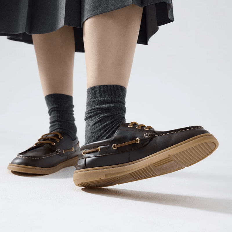 A low angle view of a person in a skirt wearing Groundies Tide barefoot boat shoes in brown leather with classic leather lacing. The left foot is rested on the heel so the outsole tread is visible.