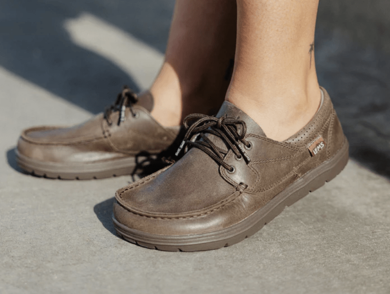 A close up of a person's feet in Lems Mariner, a barefoot boat shoe with a wide toe box and zero drop sole, in cocoa colored leather with a matching sole. The view is from the side showing the stitching detail around the toe box, and laced top