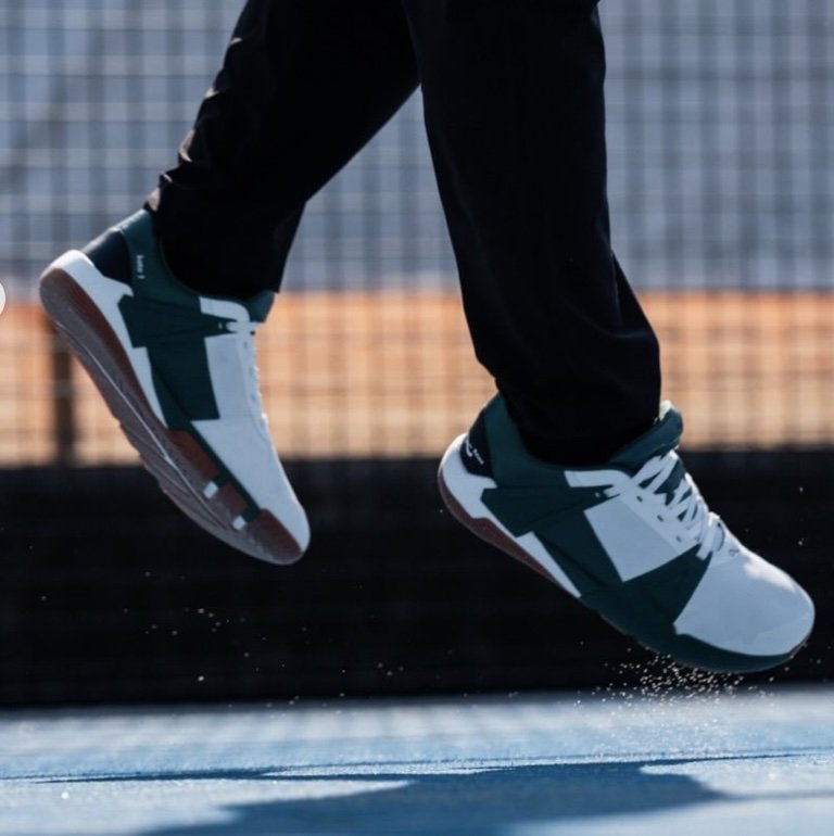 A close up of a person jumping off of a tennis court from the shins down wearing Notace white and green Koto wide toe box barefoot court tennis shoes