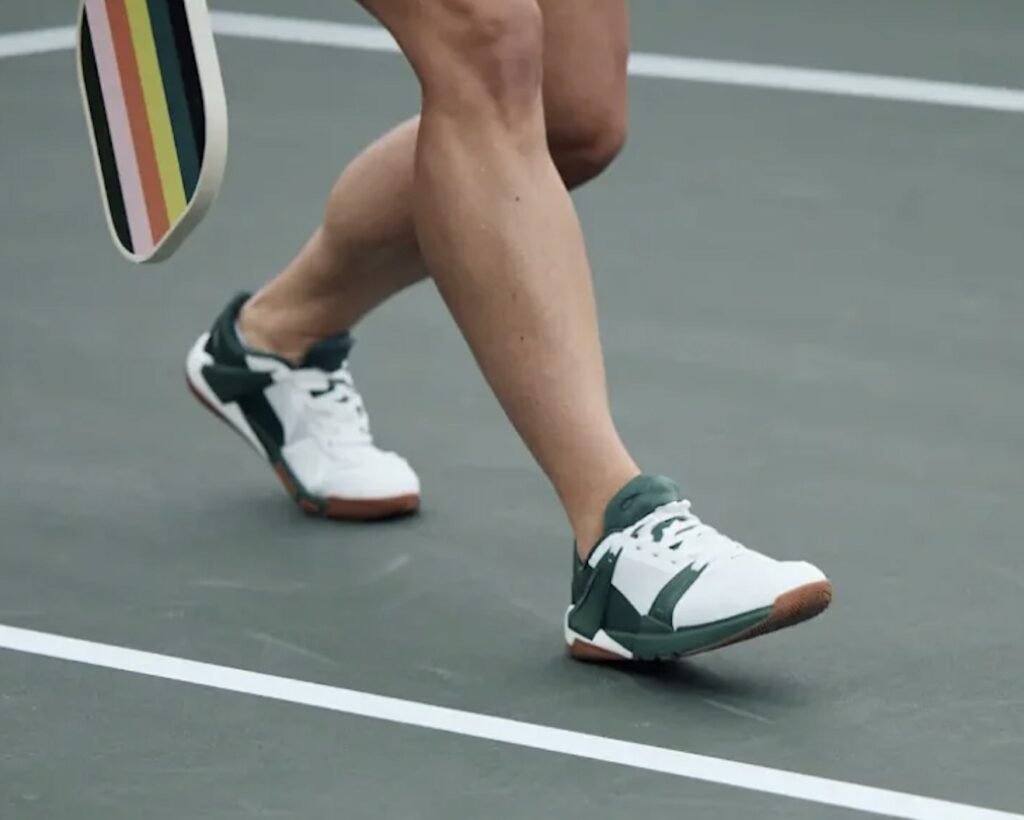 A close up view of a pair of feet on a pickleball court in mid stride holding a paddle and wearing Notace Koto wide toe box barefoot court shoes in white and green