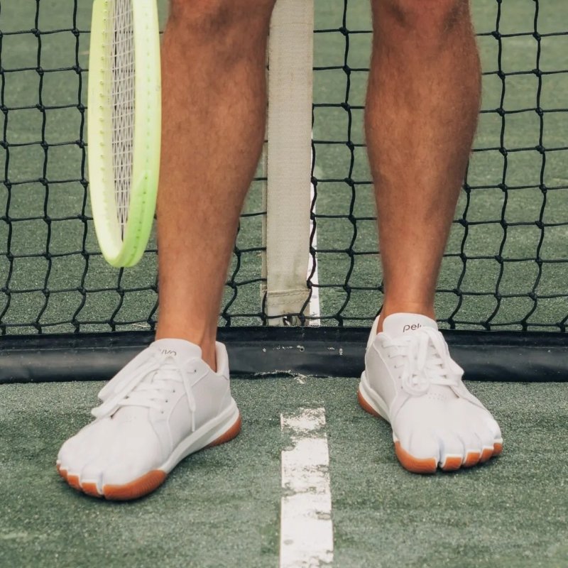 Front view of a person in Peluva Court Ad-In Five toe sneakers on a zero drop outsole designed for tennis use. The person is standing with the net behind them and a tennis raquet on their right. The five toe pockets of each shoe are clearly visible in the forefront of the photo.