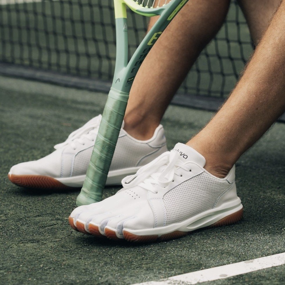 Side view of a person sitting down on a tennis court with their raquet between their feet. On their feet are a pair of Peluva Court Ad-In five toe pocket sneakers for tennis use.