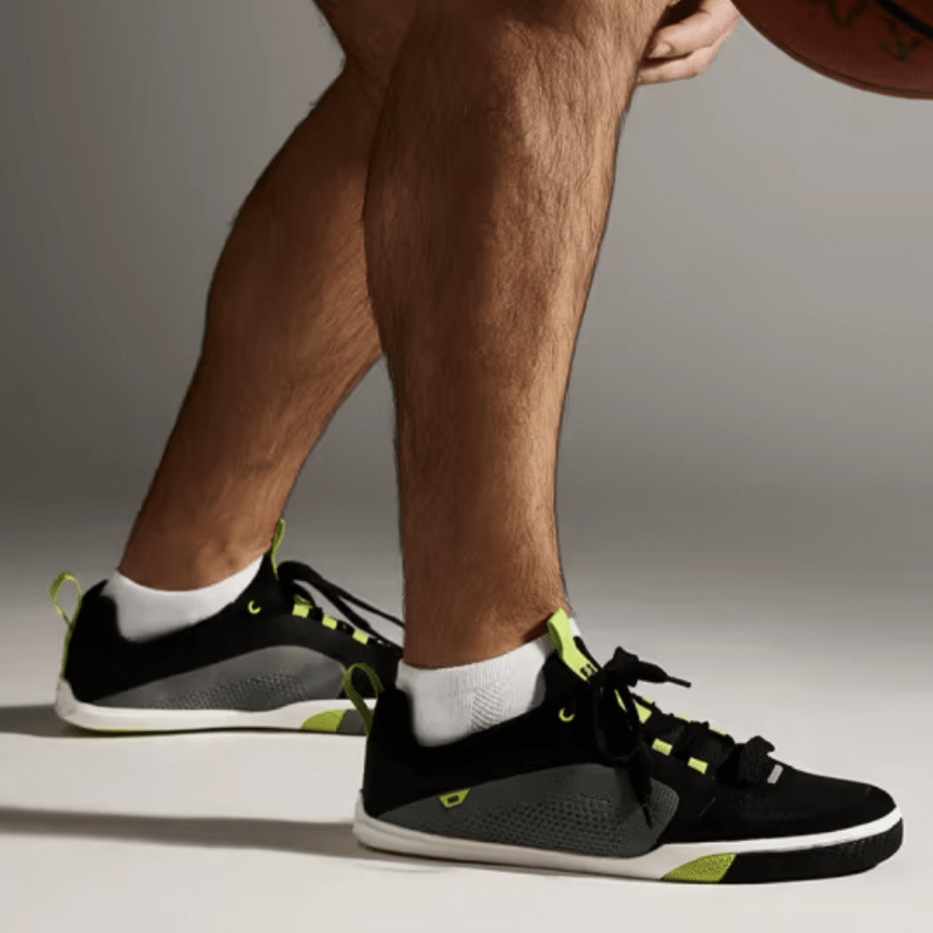 A side view of a man's legs in Pines Footwear Barefoot Court Shoes in black with gray and green accents. The zero drop wide toe box sneakers are designed for court use with a grippy outsole