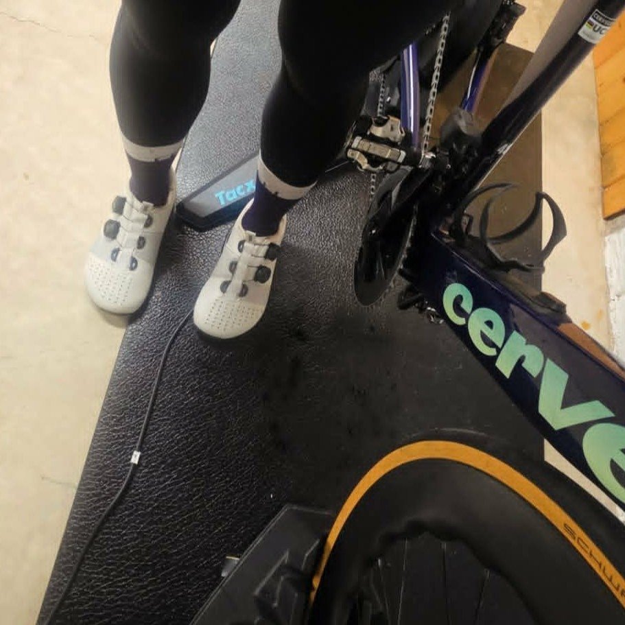 A close up of a person's feet in Strong Feet Athletics Leviathan cycling shoes with a wide toe box standing next to an indoor bike trainer