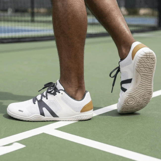 A person's feet walking on an outdoor court wearing Xero 360 Rally, a wide toe box zero drop sneaker for court sports. The person's right foot is lifted up to show the grippy outsole and wide toe box.