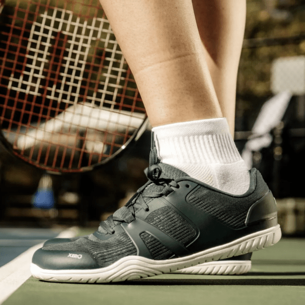 A side profile view of a person on a tennis court in Xero 360 Rally, a barefoot sneaker for court sports. There is a tennis raquet in the background.