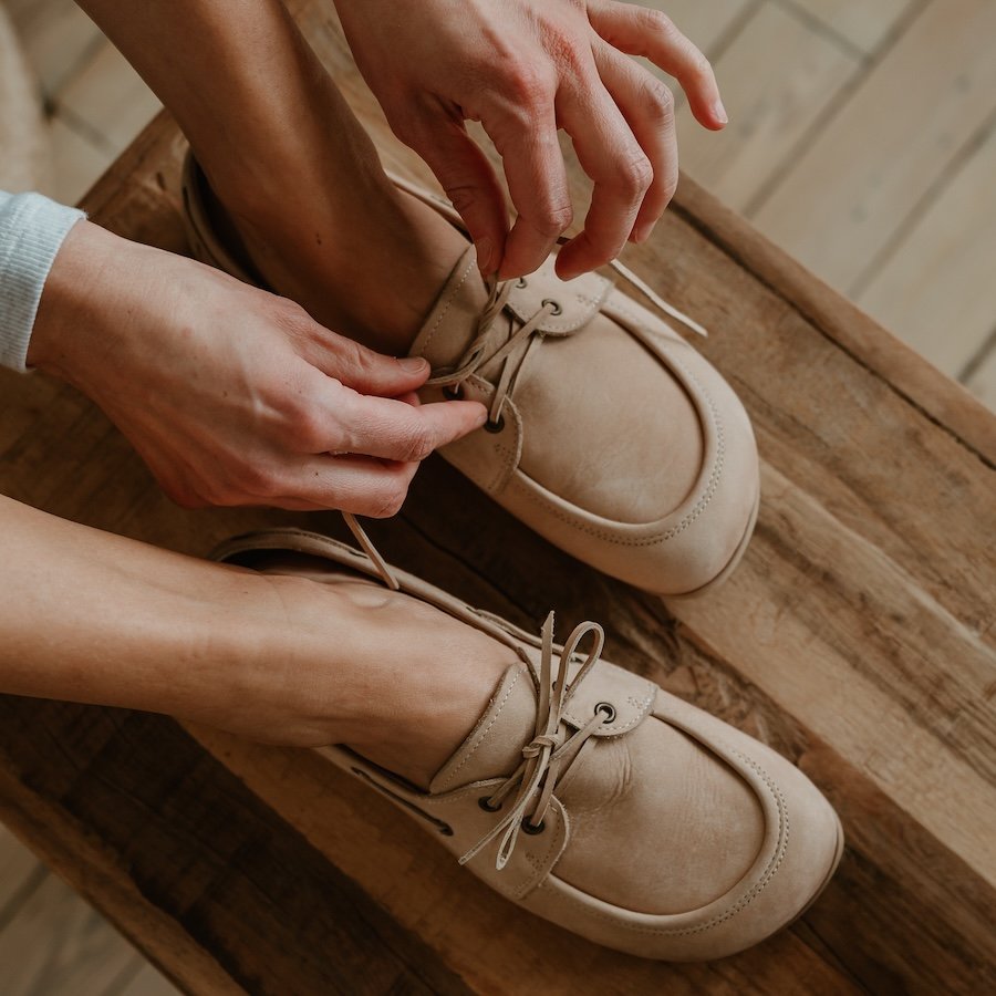 A top down view of a person in Peerko Tala boat style barefoot shoes in tan leather. They are tying the laces on the left shoe, while both feet are resting on a wooden bench